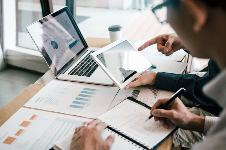 Homens com relatórios, notebook e tablets na mesa.