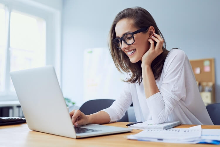 Mulher de pele clara com roupa social e óculos ao notebook, no trabalho.