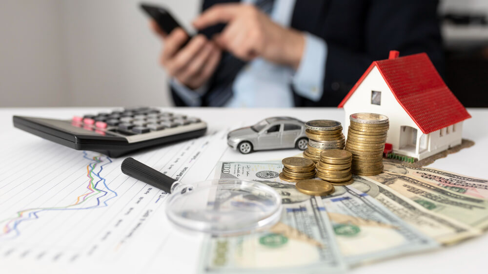 Moedas empilhadas, notas de dólar, miniaturas de casa e carro sobre uma mesa com gráfico e calculadora, representando cálculo e planejamento da margem consignável em empréstimos.